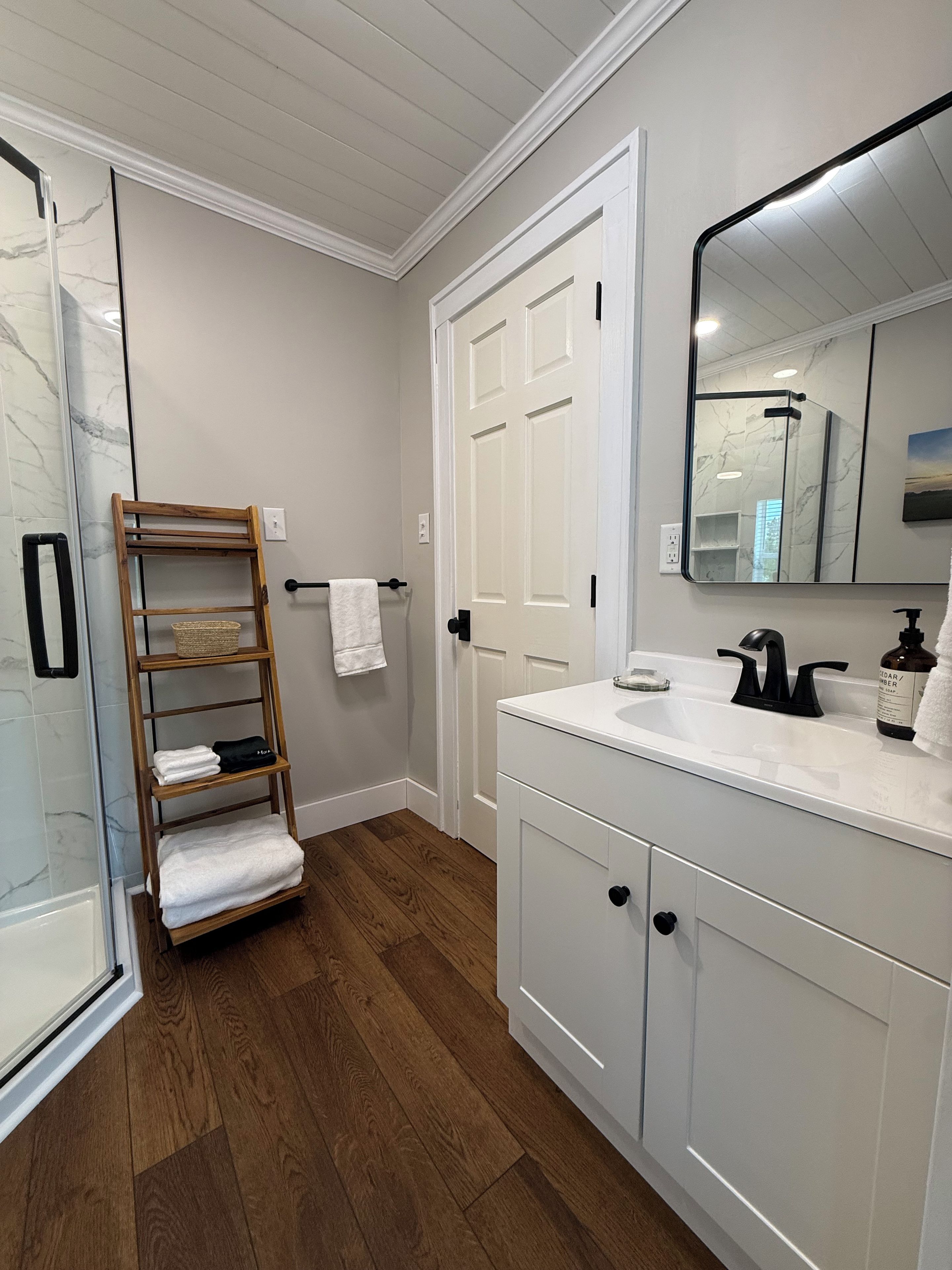 Un bagno con uno scaffale in legno, una porta bianca, uno specchio e un mobile lavabo bianco.