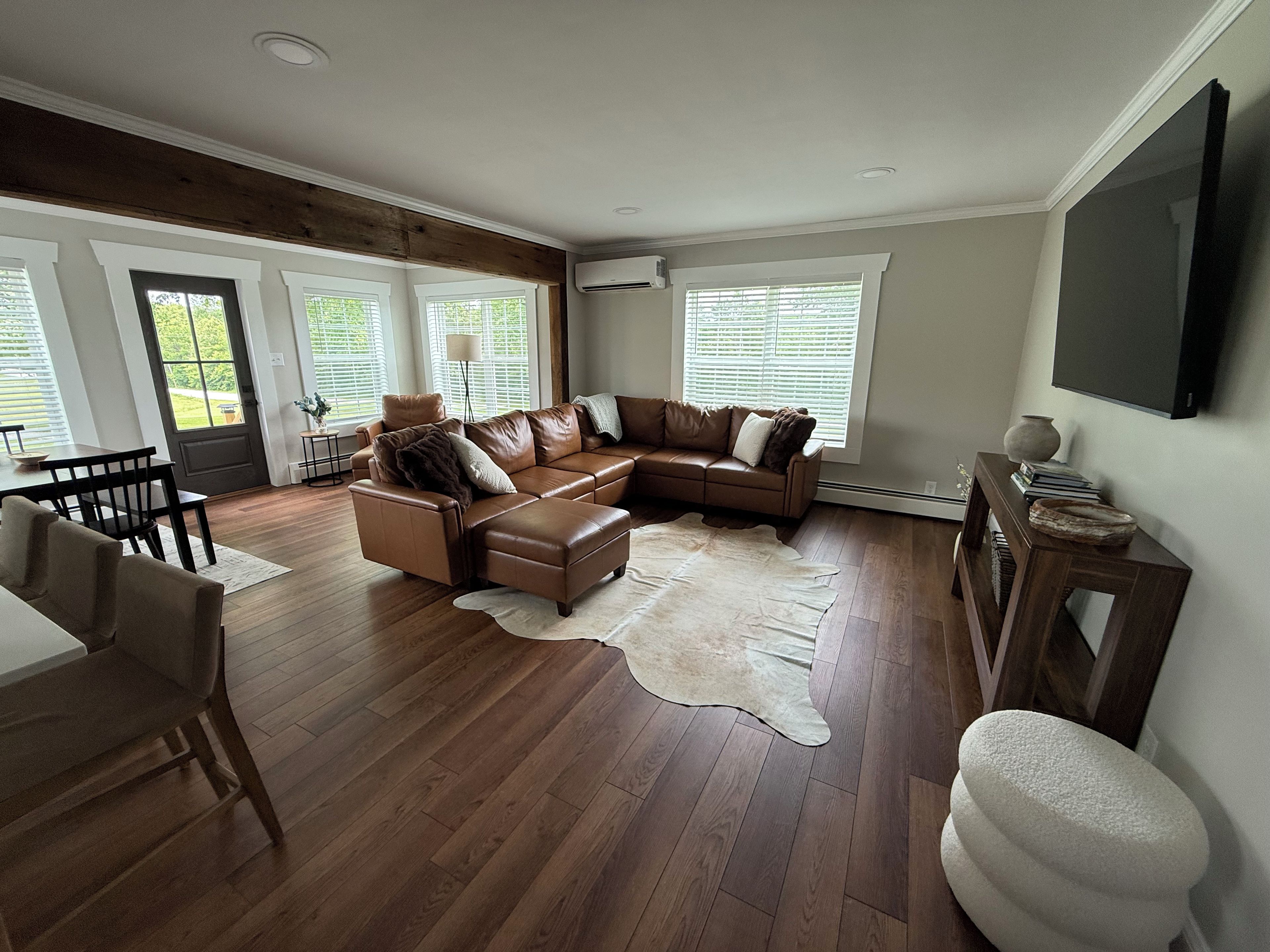 Een woonkamer met een hoekbank, een eettafel met stoelen en een groot raam met zonwering.