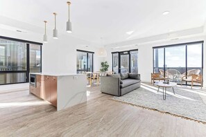 Amplia sala de estar con sofá, mesa de comedor, sillas, ventanas grandes y una zona de cocina.