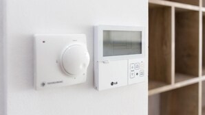 Ein Thermostat mit digitaler Anzeige und Bedienelementen, an einer weißen Wand angebracht.