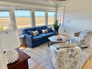 Un salon avec un canapé bleu, des fauteuils à motifs, une table basse et une vue sur la plage.