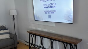 Una sala de estar con un televisor de pantalla plana montado en la pared, un sofá gris, una lámpara de pie, una mesa de centro de madera y una consola con estructura metálica.