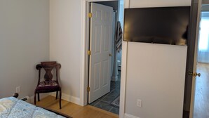 Un dormitorio con cama, una silla, una puerta y televisor montado en la pared.