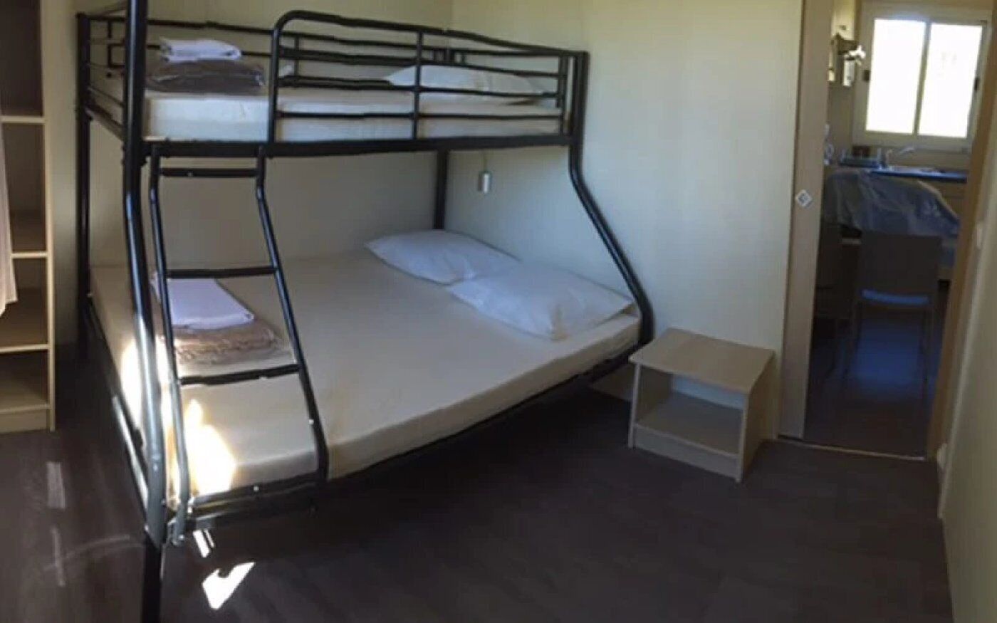 Uma cama beliche com uma mesinha e uma prateleira, uma porta para o banheiro e uma janela.