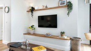 Een woonkamer met een flatscreen televisie, houten planken met planten, een bijzettafeltje met boeken en een cocktailshaker, en een rieten mand.