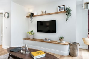 Una sala de estar con un televisor de pantalla plana, estantes de madera con plantas, una mesa de centro con libros y un agitador de cócteles, y una cesta de mimbre.