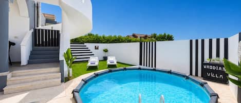 A modern swimming pool with a white deck, a set of stairs leading to a covered area, and a sign reading 'RHODIANA VILLA'.