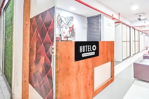 A hotel reception desk with a 'HOTEL' sign, a wall-mounted artwork, and a red railing along the corridor.