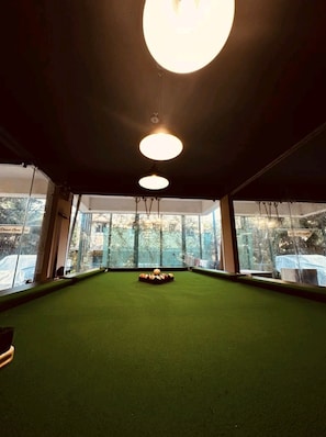 Una mesa de billar con superficie de fieltro verde, rodeada de amplias ventanas y tres lámparas colgantes.