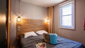 Quarto com cabeceira de madeira, duas luminárias de cabeceira, uma janela com persianas, cama com cobertura escura e uma revista na cama.
