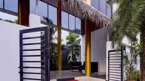 A modern building entrance with a metal gate, a red carpet, and a staircase leading to a covered area with outdoor seating. - Deluxe Vanam Resort (Vanur)