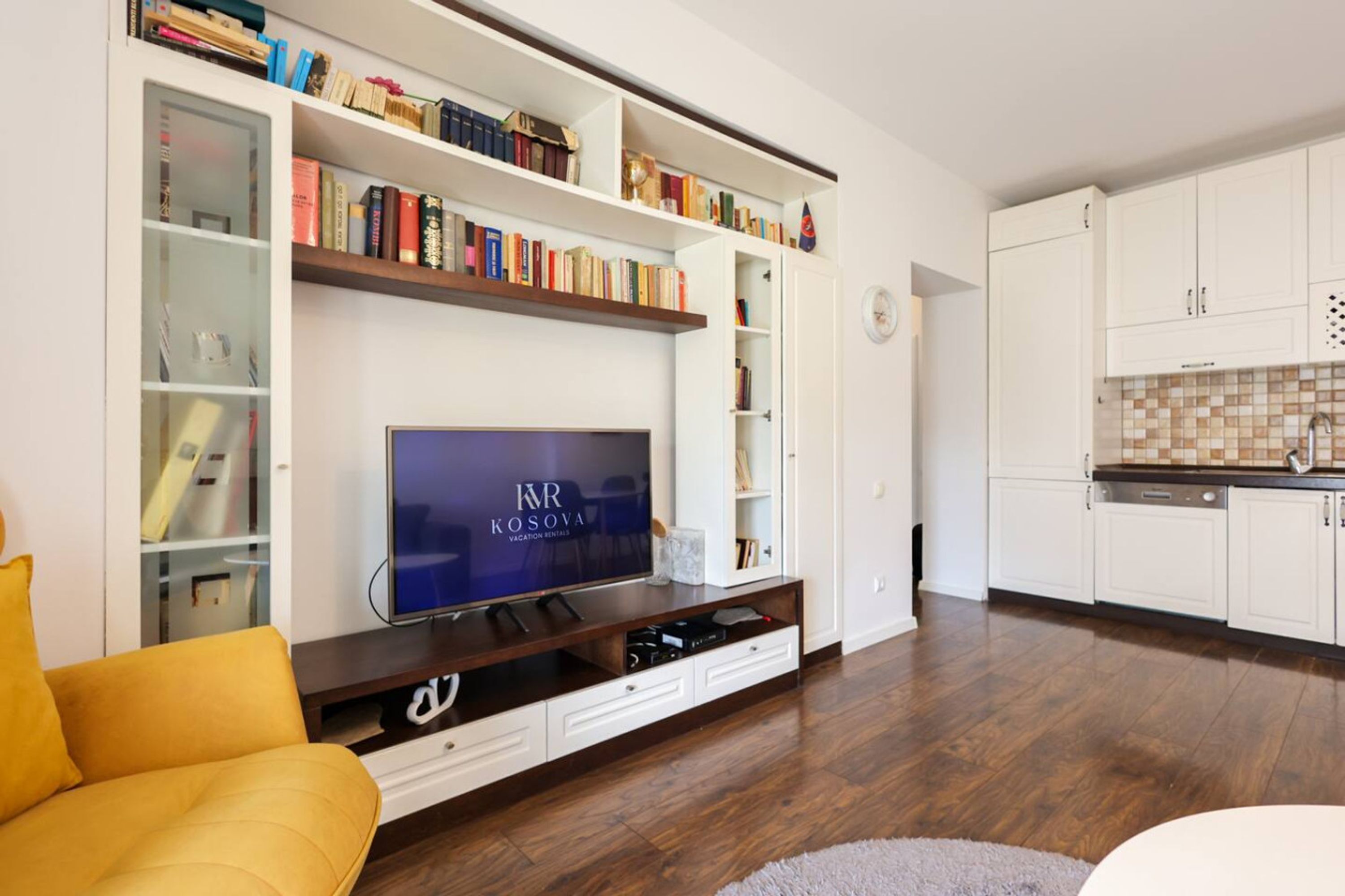 A modern living room with a flat-screen TV, a bookshelf, a sofa, and a kitchen area.