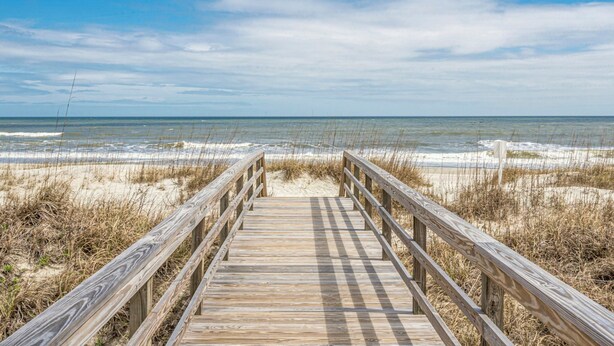 A wooden boardwalk leading to a beach with sand and grass.