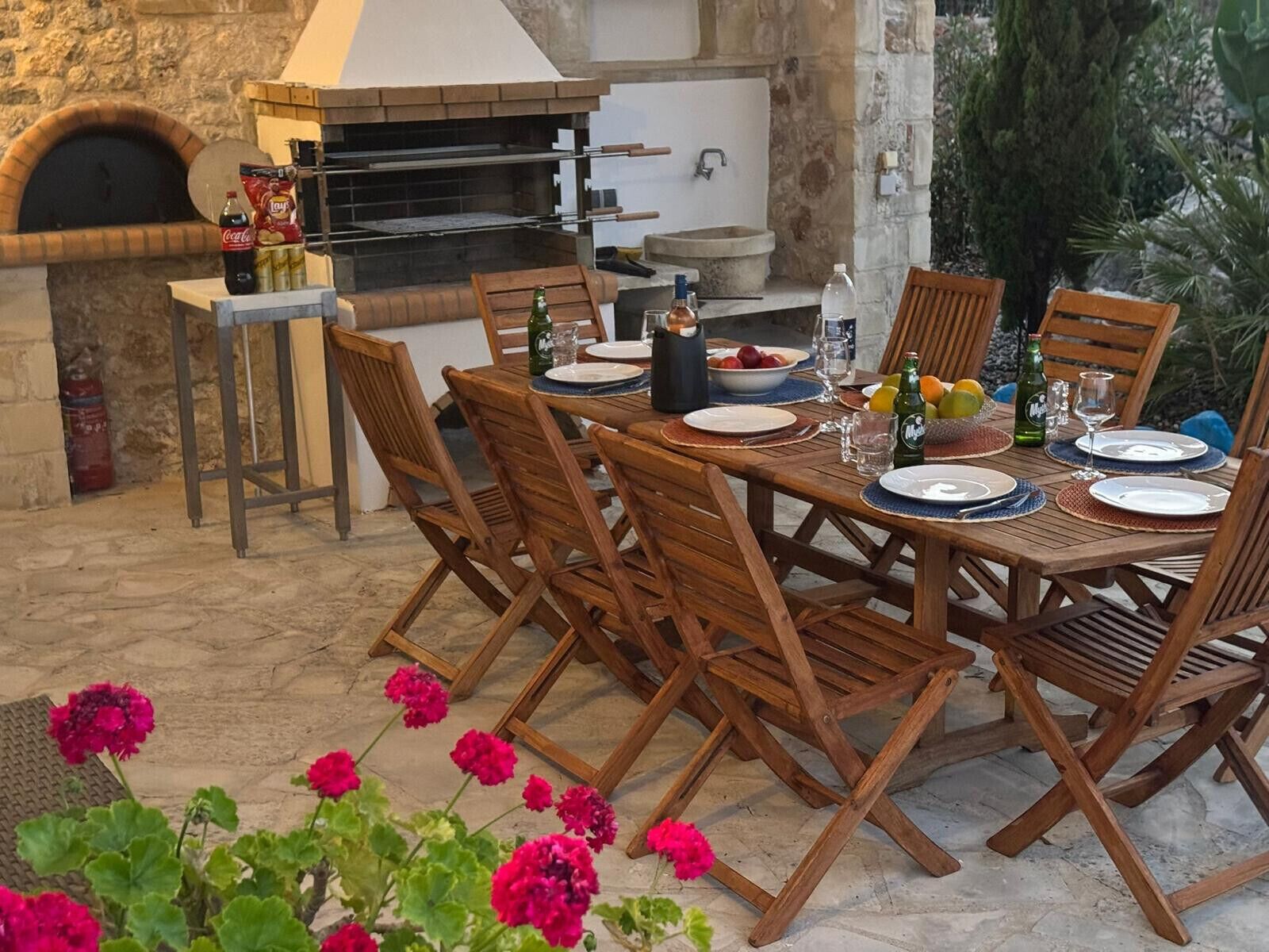 An outdoor dining area with a brick oven, wooden chairs, and a table set for four.