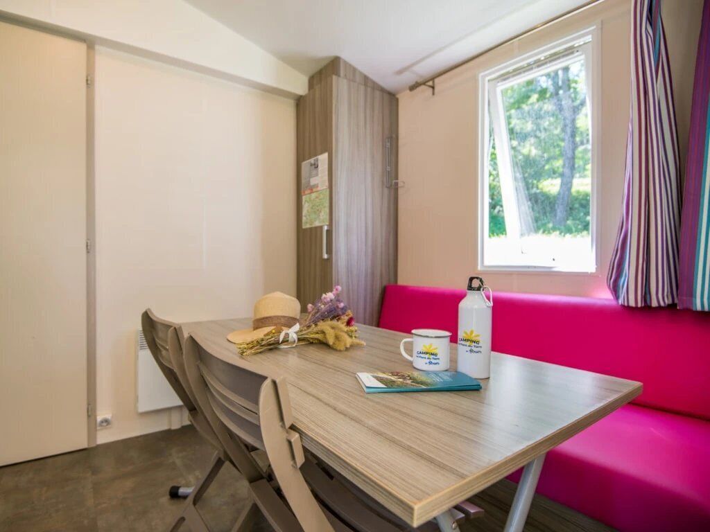 Une pièce avec une table en bois, un canapé rose, une fenêtre avec des rideaux et une bouteille d’eau.