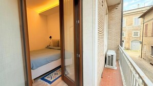 Een slaapkamer met een bed, een schuifdeur, een kleedje en een balkon met een hekwerk en een gebouw op de achtergrond.
