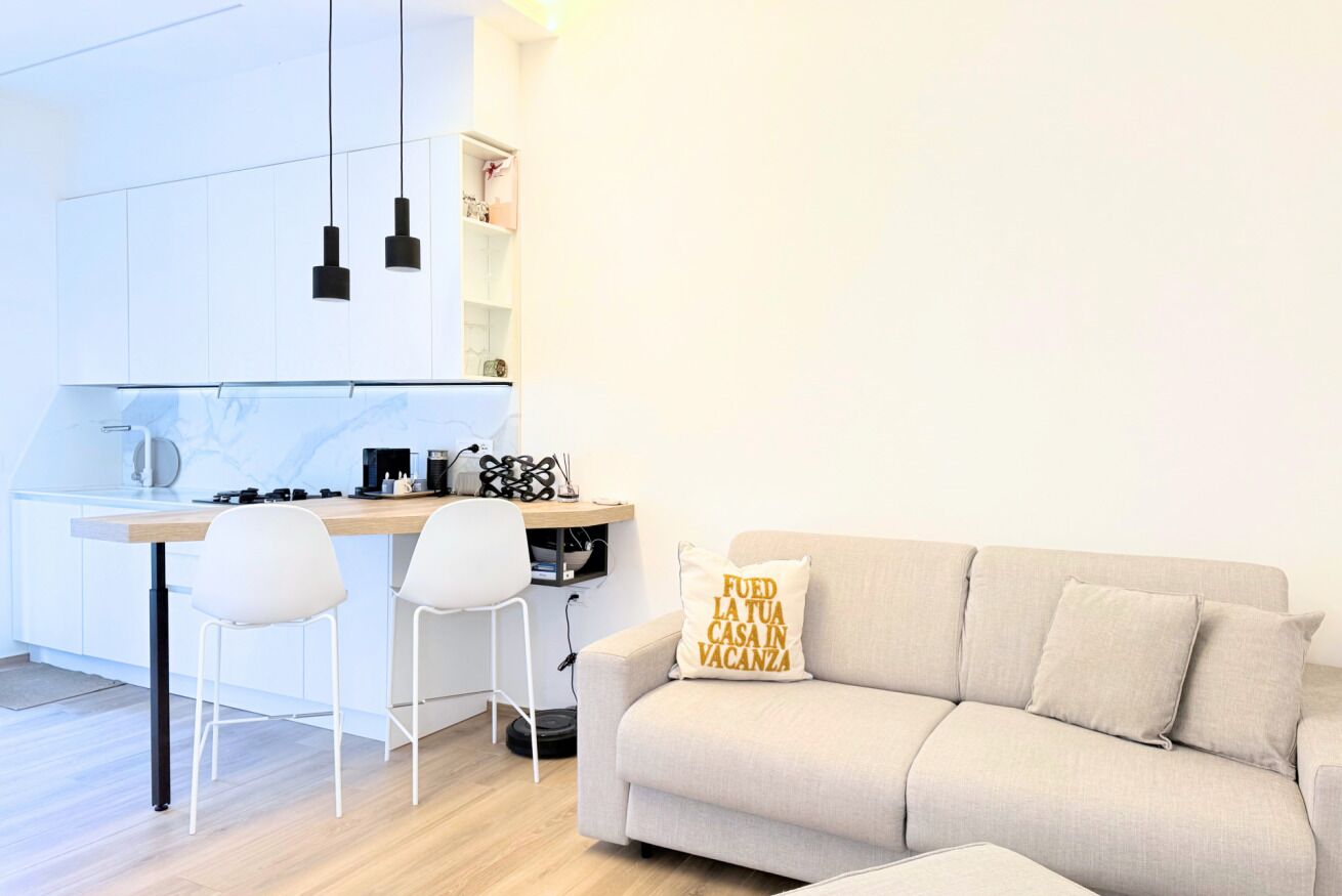 A modern kitchen with white cabinets, a dining area with two chairs, and a sofa with a cushion.