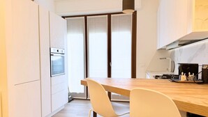 Een moderne keuken met een houten tafel, witte stoelen, een inbouwoven en een groot raam met gordijnen.
