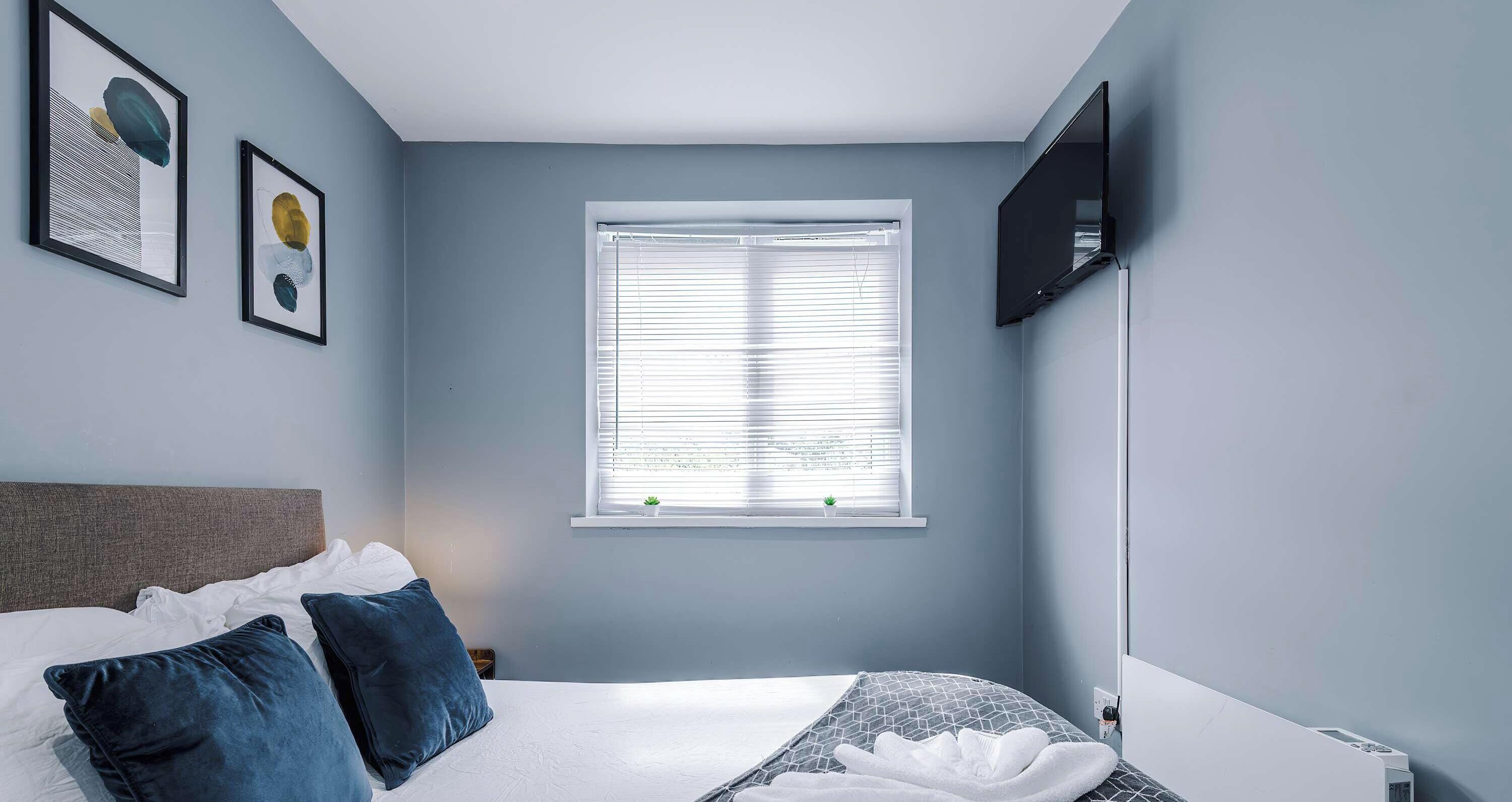 A bedroom with a bed, pillows, a window with blinds, a wall-mounted TV, and framed artwork.