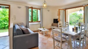 A room with a sofa, dining table, chairs, and a TV. There are windows with a view of greenery outside. - Pascaline, Holiday Cottage for 4 People (Le Bar-sur-Loup)