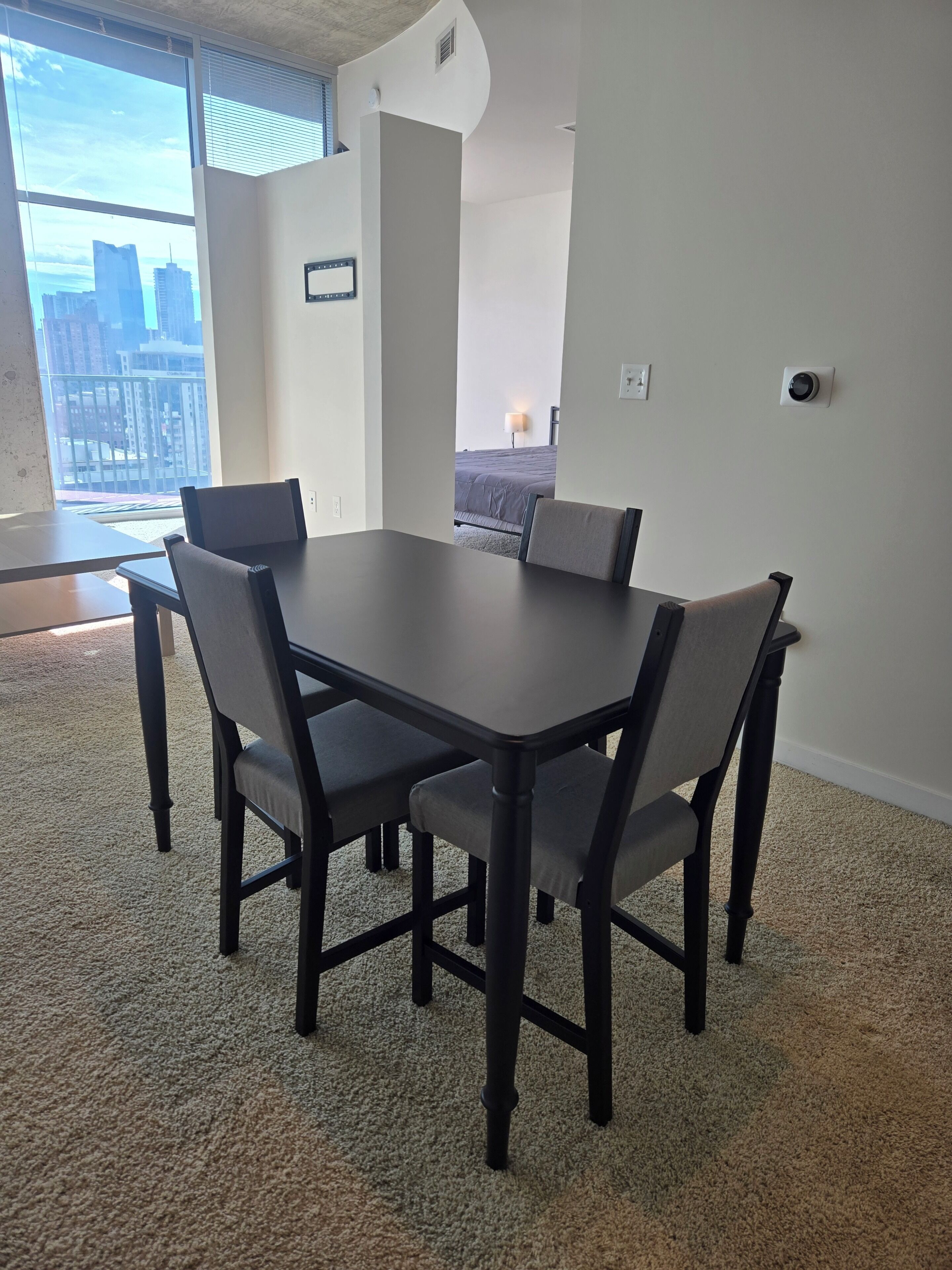 A modern dining area with a rectangular table and four chairs, carpeted floor, and a view of a cityscape through large windows.