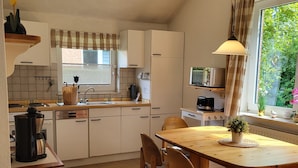 A kitchen with white cabinets, a wooden dining table, and a window with curtains. - Haus Teelke vacation home in an idyllic location directly on the river (Papenburg)