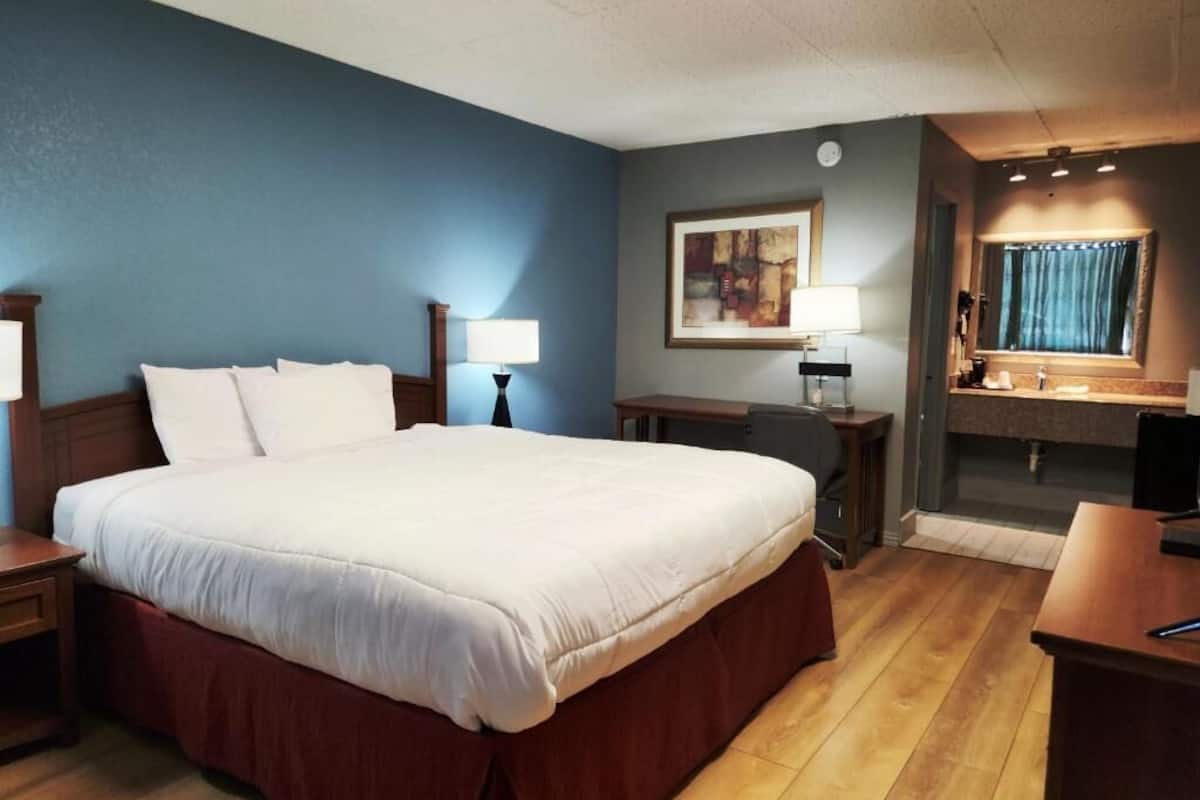 A hotel room with a large bed, bedside tables, a desk, a chair, a window with curtains, and a painting on the wall.