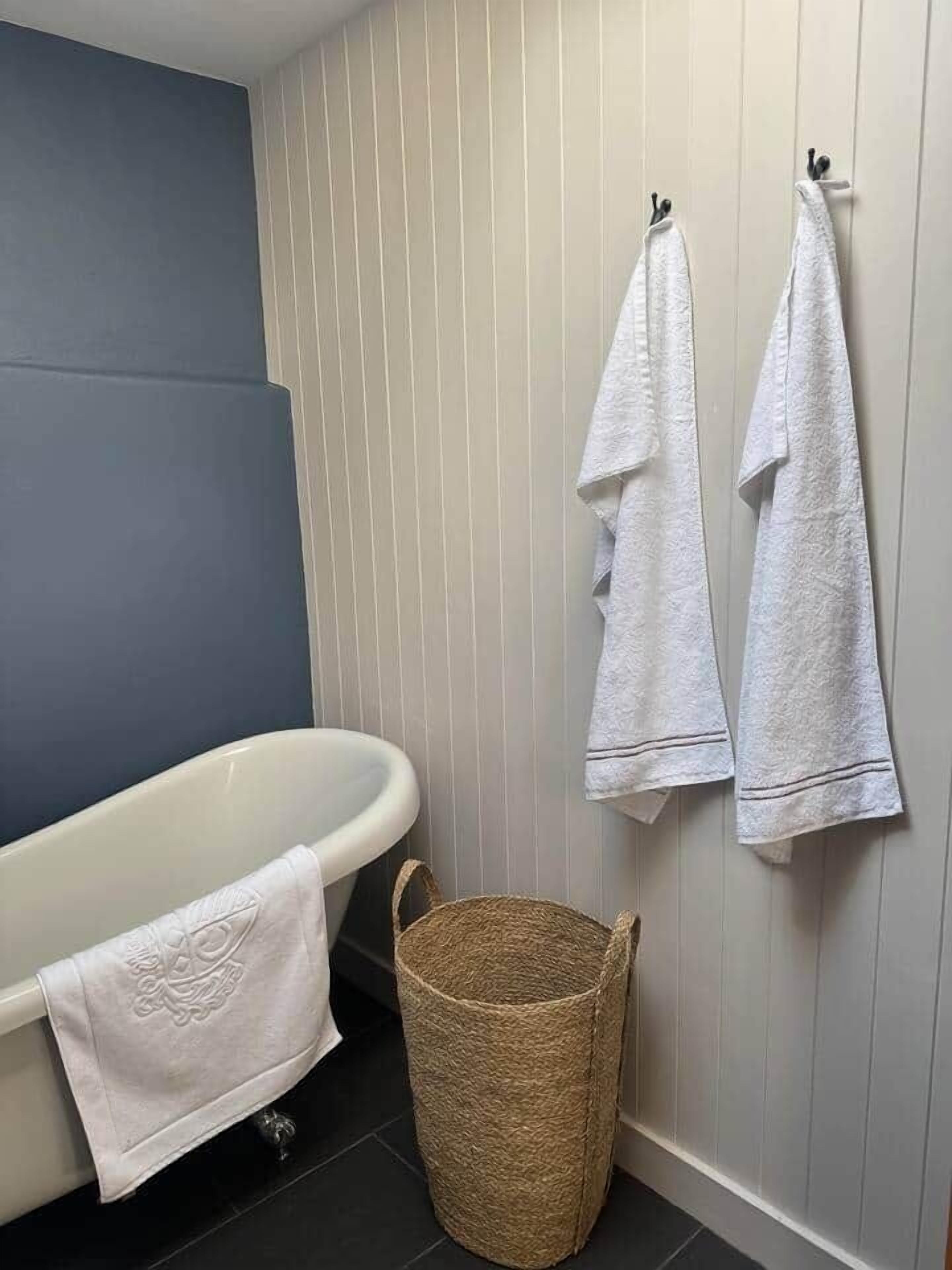 Ein Badezimmer mit einer weißen Badewanne, zwei weißen Handtüchern, die an Haken hängen, und einem gewebten Korb auf dem Boden.