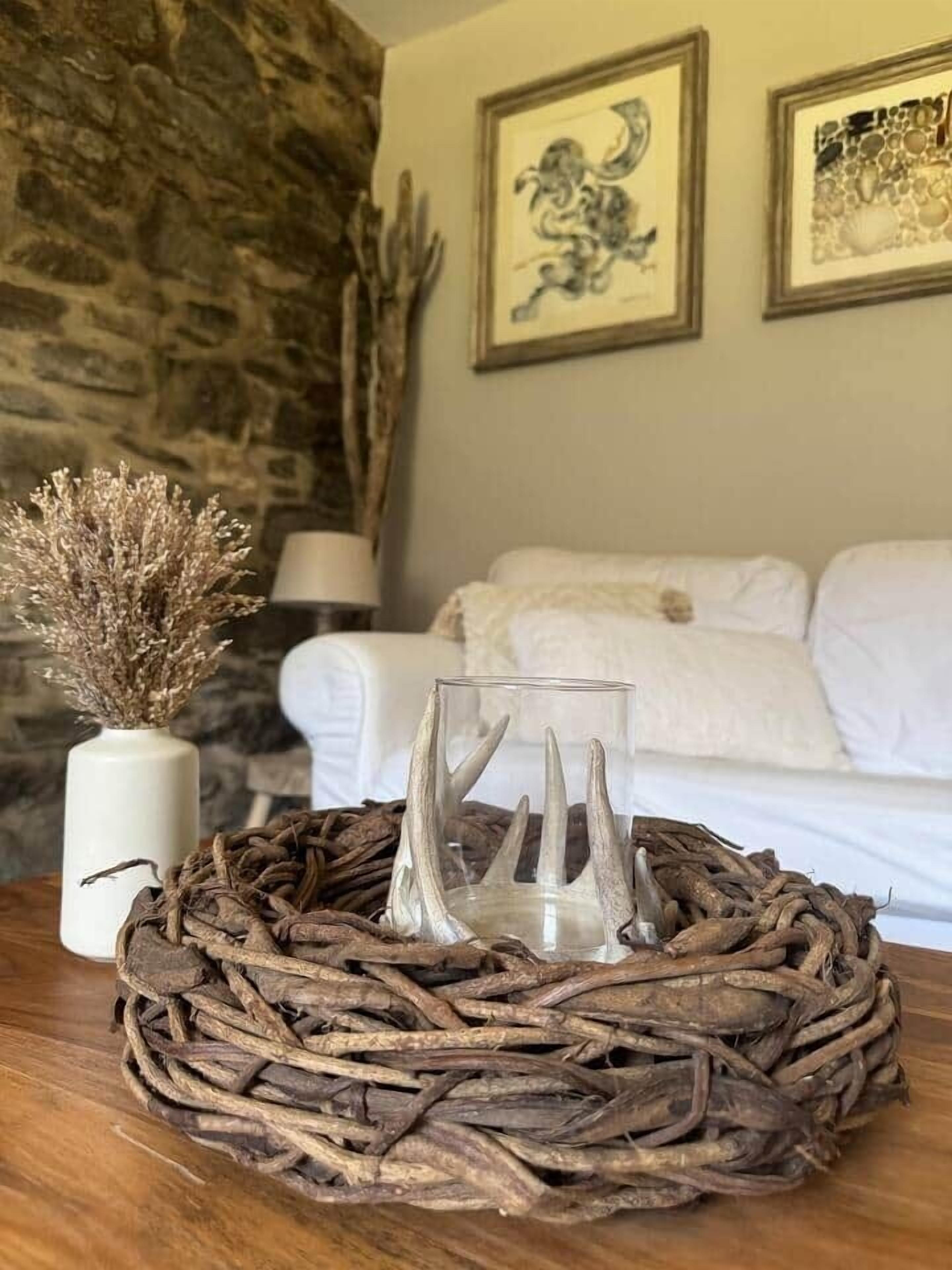 Un décor rustique avec un panier tressé posé sur une table en bois, un vase contenant des plantes séchées, un vase en verre, et un lit avec du linge de maison blanc en arrière-plan.