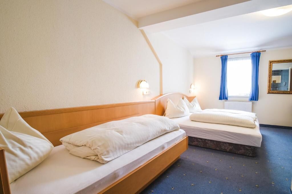 Una camera d'albergo con due letti singoli, una finestra con tende blu, uno specchio e un punto luce a parete.