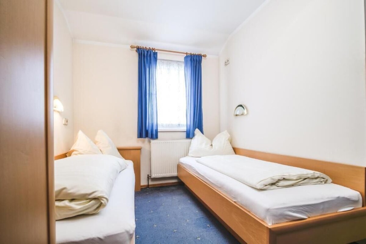 A small hotel room with two single beds, a window with blue curtains, a radiator, and a wall-mounted light fixture.