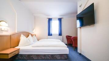A hotel room with a bed, bedside table, lamp, TV, window with curtains, and a red chair.