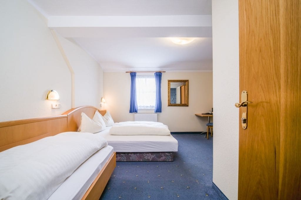 A hotel room with two beds, a desk, a chair, a window with curtains, and a door.