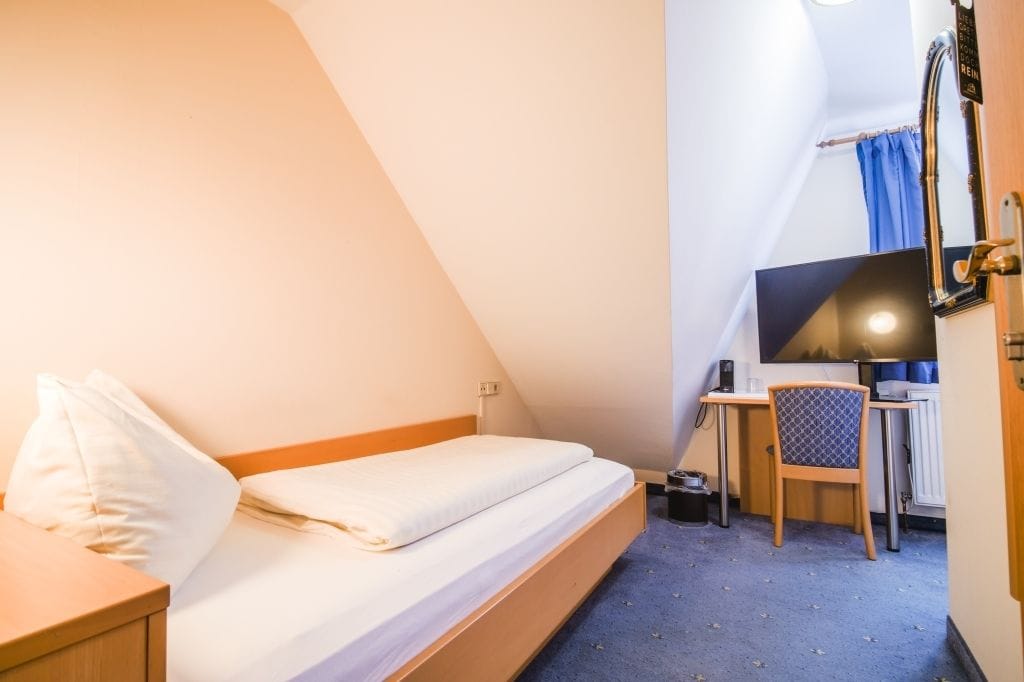 Una piccola stanza triangolare con un letto, una scrivania, una sedia e una televisione.