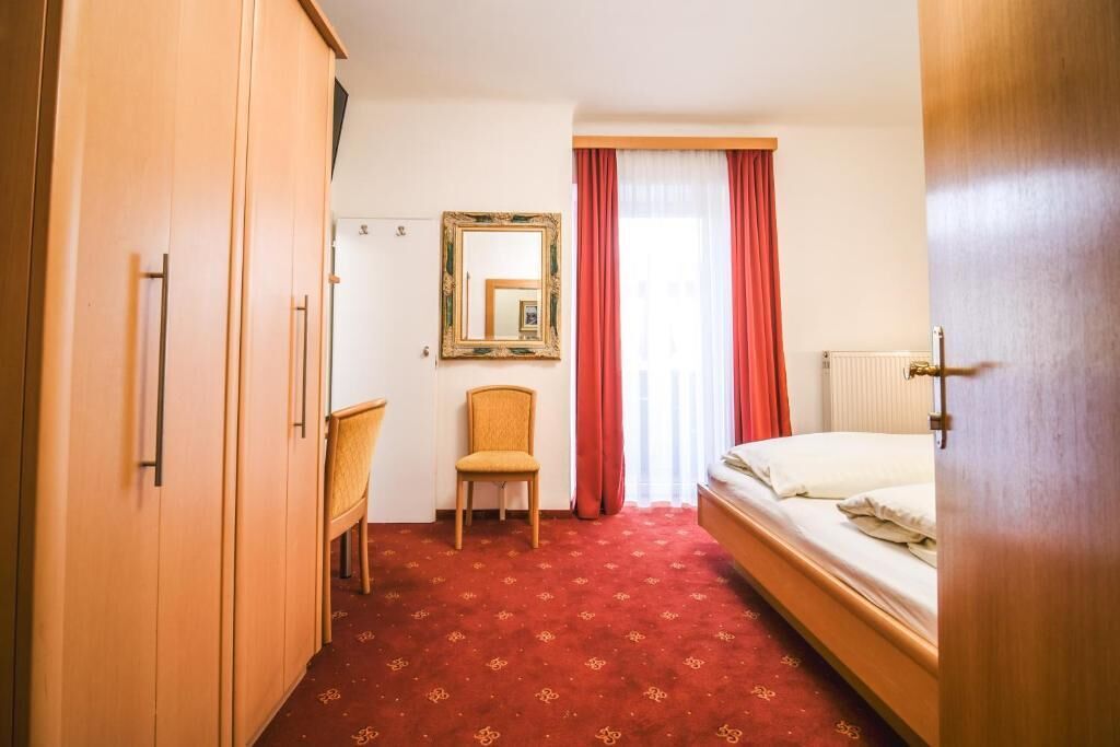 Une chambre d’hôtel avec un lit, une chaise, une armoire, un miroir et une fenêtre avec des rideaux.