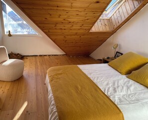 A bedroom with a sloped ceiling, wooden flooring, a bed with a white and mustard-colored bedspread, and a window with a view of trees. - Apartment in Serre Chevalier, near the slopes - 3 bedrooms - sleeps 6 (Le Monêtier-les-Bains)