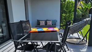 A covered patio with a table and chairs, a cushioned seating area, and a view of greenery. - Spacious, bright villa ideal for a stay in Mimizan with private pool (MIMIZAN)