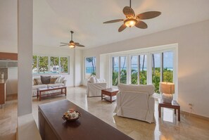 Un vaste salon doté d’une grande fenêtre donnant sur une plage, d’un ventilateur de plafond et d’un coin repas avec une table en bois.