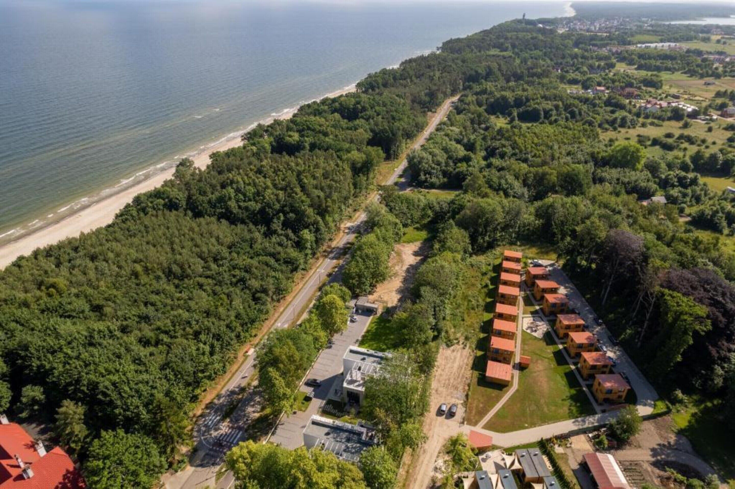 An aerial view of a coastal area with a beach, dense forest, and a row of red-roofed buildings.