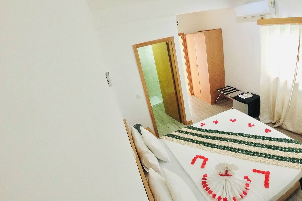 A hotel room with a bed, a small table, a wardrobe, and a door leading to another room.