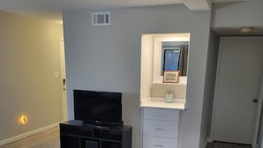 A living room with a flat-screen TV, a white cabinet with a clock, and a brown ottoman. - 2-bedroom condo in gorgeous Calabash with cool AC breeze (Calabash)
