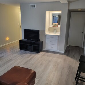 A living room with a flat-screen TV, a white cabinet with a clock, and a brown ottoman. - 2-bedroom condo in gorgeous Calabash with cool AC breeze (Calabash)