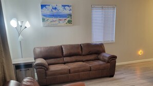 A living room with a brown leather sofa, a coffee table, a floor lamp, a painting, and a window with blinds. - 2-bedroom condo in gorgeous Calabash with cool AC breeze (Calabash)