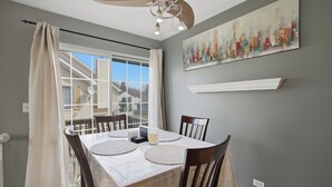 Un comedor con una mesa dispuesta para cuatro personas, un ventilador de techo y un cuadro con vista a la ciudad.