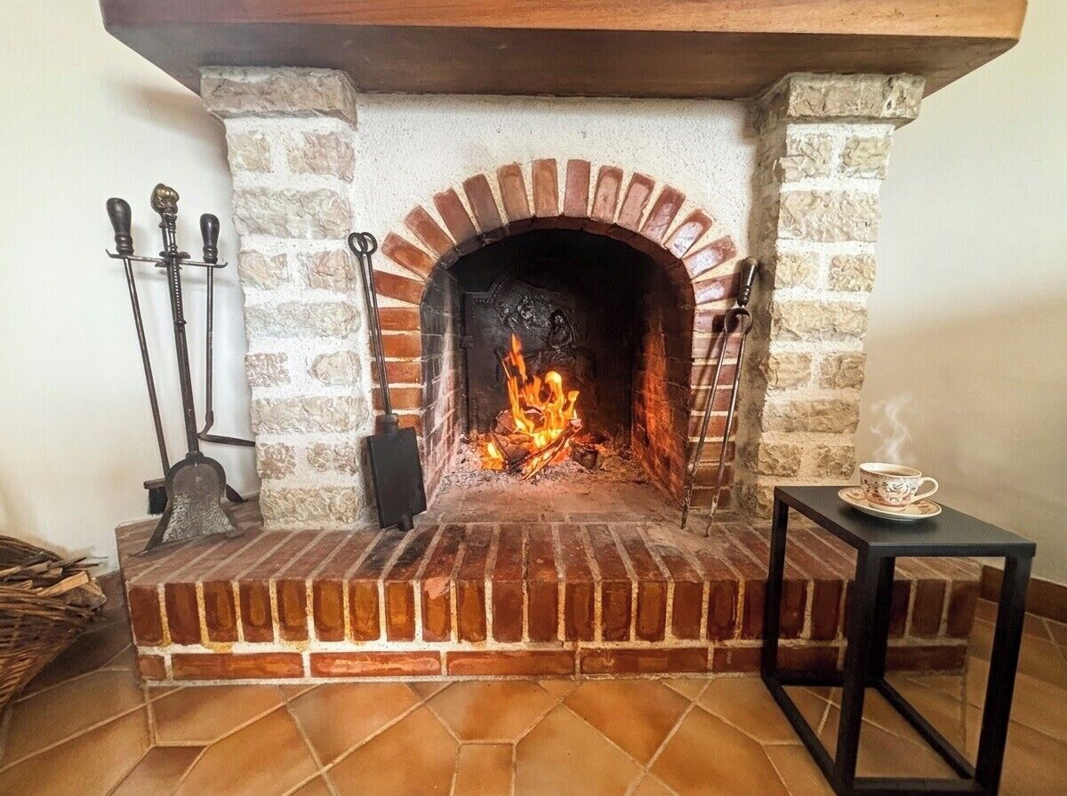 A brick fireplace with a fire, a metal table with a cup, and a basket of wood.