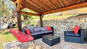 An outdoor patio with a stone wall, a wooden pergola, a red chair, a grey sofa, and a black sofa with red cushions. - The Olive Grove - Sea View (Saint-André-de-la-Roche)