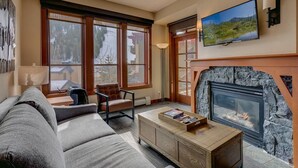 A living room with a stone fireplace, a television mounted above it, and a large window with a view of a snowy landscape. - Palisades View One Bedroom (Olympic Valley)