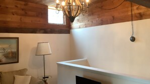 Une pièce avec des poutres apparentes, un lustre, une cheminée blanche, une lampe et un tableau au mur.