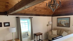 Une pièce avec un plafond en bois, un lustre, une fenêtre avec des rideaux, un canapé, une lampe, un tableau et une petite table.