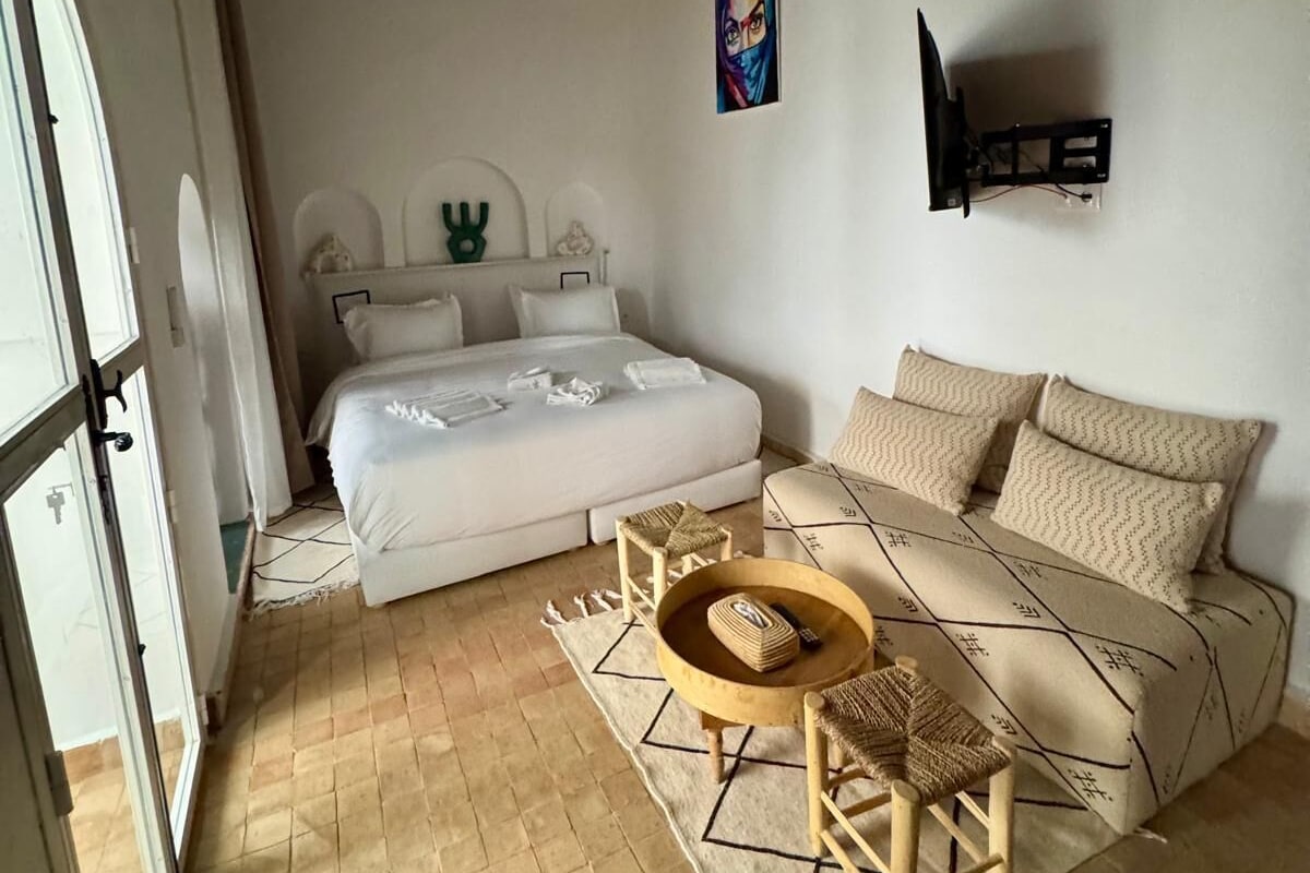 A bedroom with a bed, a sofa, a small wooden table, a hanging light fixture, and a painting on the wall.